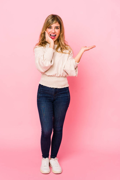 Young blonde caucasian woman standing over a pink background holds copy space on a palm, keep hand over cheek. Amazed and delighted. - Foto, Imagem