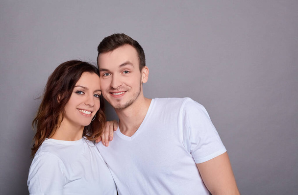 Portrait of a Caucasian lovely couple - a man with blue eyes hugs his charming, sweet woman, and they both look at the camera, standing against a gray background. Copy space. - Photo, Image