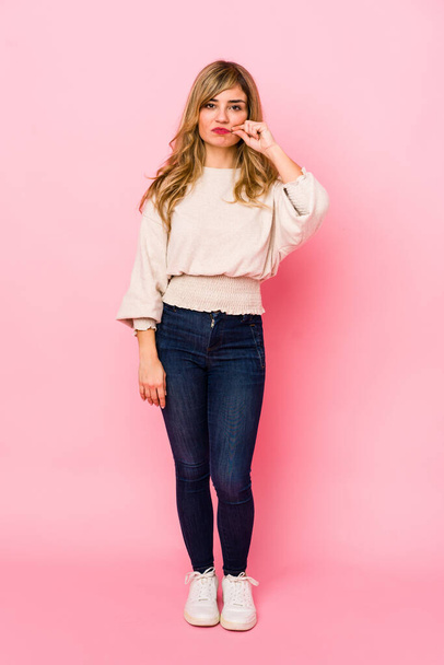 Young blonde caucasian woman standing over a pink background with fingers on lips keeping a secret. - Фото, зображення