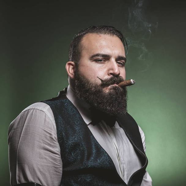 Ένας άντρας με γενειάδα καπνίζει πούρο, κοιτάζει την κάμερα..  - Φωτογραφία, εικόνα