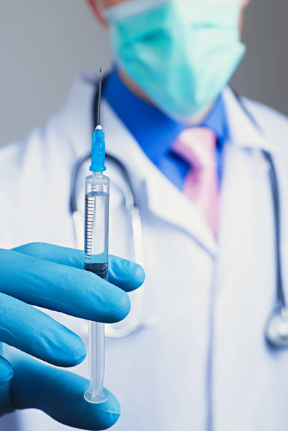 Close up view of medical syringe in rubber blue glove dressed hand, man chest in white medical robe with stethoscope on a blurred background - Zdjęcie, obraz