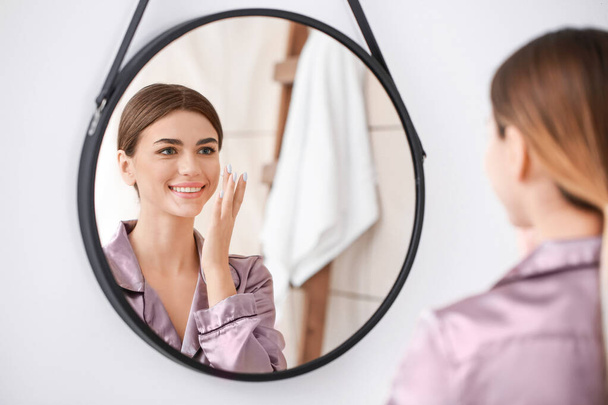Красивая молодая женщина наносит крем на лицо возле зеркала в ванной комнате
 - Фото, изображение