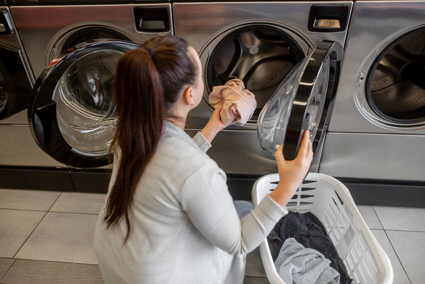 hübsche junge Frau, die ein paar schmutzige Kleider in eine Waschmaschine in einer Wäscherei bringt, Wäschewaschen - Foto, Bild