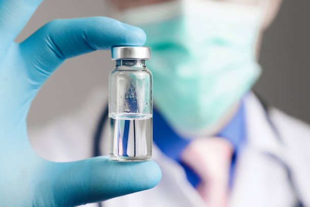 Cerca de la mano del médico en guante de látex azul y máscara de gasa médica con ampolla de vidrio. Concepto de salud y vacunación
 - Foto, Imagen