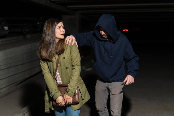 Pelokas nuori latino nainen katselee takaisin ryöstäjä uhkaa häntä veitsellä parkkipaikalla
 - Valokuva, kuva