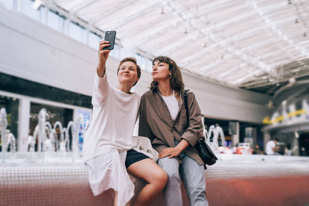 Two girls take a selfie in the mall, a fountain in the background - Zdjęcie, obraz