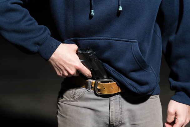 Midsection of male robber removing gun from jeans while standing at parking lot - 写真・画像