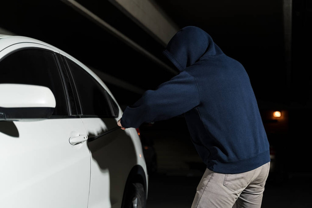 Ladrão com capuz tentando quebrar a porta do carro aberto com pé de cabra no estacionamento escuro
 - Foto, Imagem