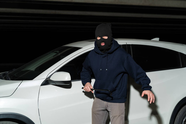 Portrait of male robber in ski mask holding crowbar while standing against car in parking lot - Foto, imagen