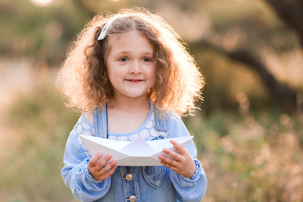 Smiling baby girl 3-4 year old holding paper origami boat outdoors. Childhood. Summer time.  - Photo, image