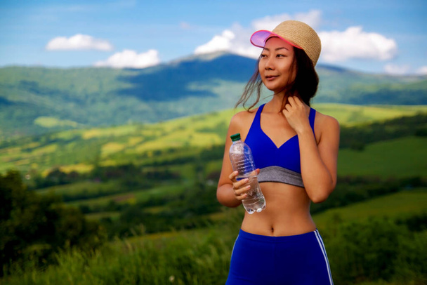 Fitness όμορφη Ασιάτισσα γυναίκα πόσιμο νερό και εφίδρωση μετά την άσκηση το καλοκαίρι ζεστή μέρα στο λόφο. Γυναίκα αθλήτρια μετά την προπόνηση. - Φωτογραφία, εικόνα