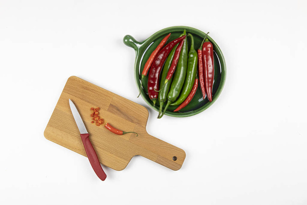 Green and red hot chilli peppers on green plate, sliced pepper on a wooden board with knife, white background, chopping board, flat lay. Seasoning - Photo, Image