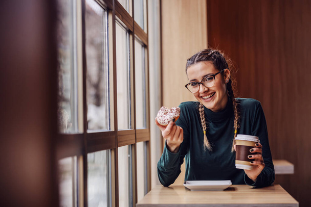 Mujer sonriente sentada en la cafetería, sosteniendo rosquilla y taza de café en el descanso entre clases
. - Foto, imagen