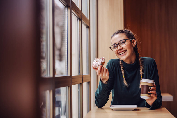 Mulher sorridente sentada no café, segurando donut e xícara descartável com café na pausa entre as aulas
. - Foto, Imagem