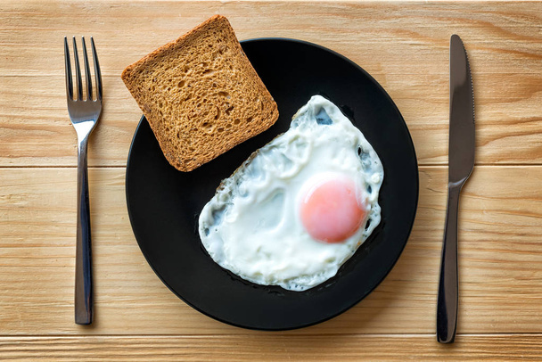 Fried egg close up in a black plate on a wooden table with toast bread and utensils. Breakfast food. Sunny side soft fried egg - Foto, afbeelding