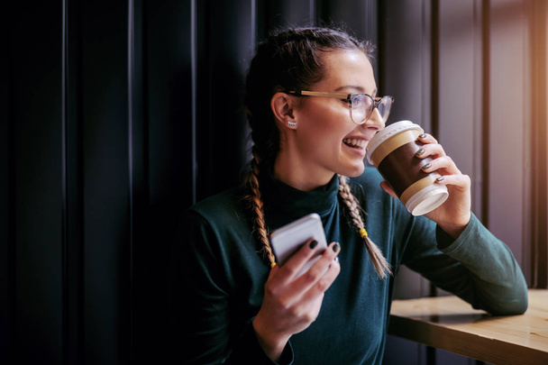 Primer plano de la mujer sonriente sentada en la cafetería junto a la ventana, bebiendo café y sosteniendo el teléfono inteligente mientras mira por la ventana
. - Foto, Imagen