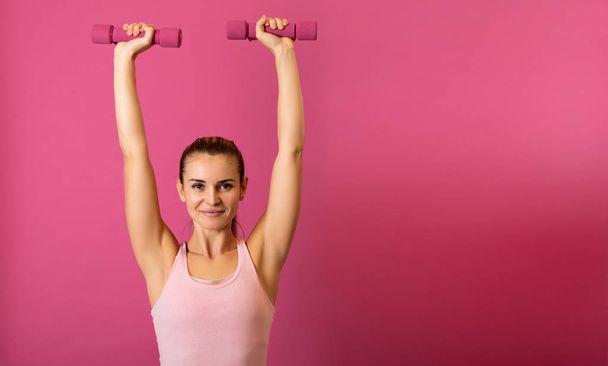 Attractive fitness woman holding dumbells on pink background - 写真・画像