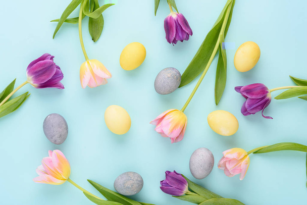 horní pohled Velikonoční pozadí fialových a růžových tulipánů, pastelově žlutá, modrá vejce na modrém pozadí, slavnostní kompozice koncept - Fotografie, Obrázek