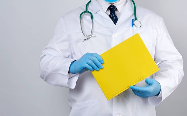 ボタン付きの白いコートの男性医師青の滅菌手袋を着用し白いスタジオの背景に黄色い紙のノートを手に - 写真・画像
