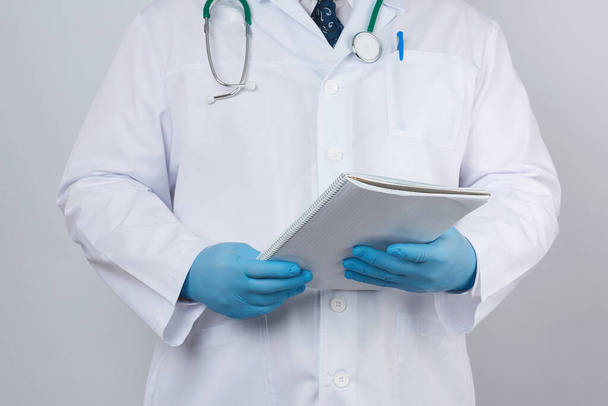 male doctor in a white coat with buttons, wearing blue sterile gloves, holding a paper notebook in his hand on a white studio background - Фото, изображение