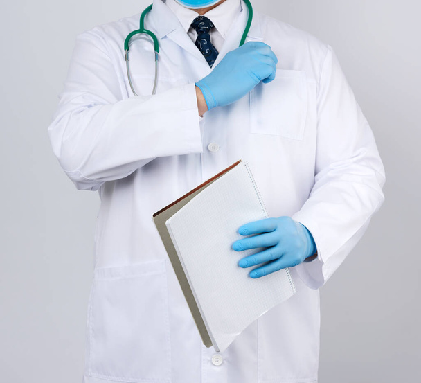 male doctor in a white coat with buttons, wearing blue sterile gloves, holding a paper notebook in his hand on a white studio background - Foto, Imagem