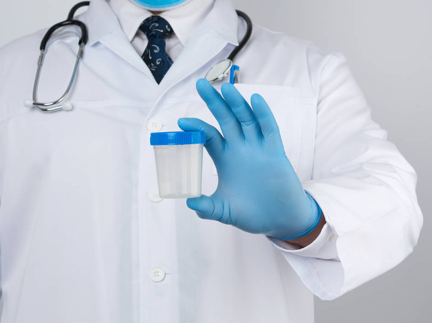 médico do sexo masculino em um casaco branco e gravata fica e mantém um recipiente de plástico para amostra de urina, vestindo luvas médicas estéreis azuis
 - Foto, Imagem