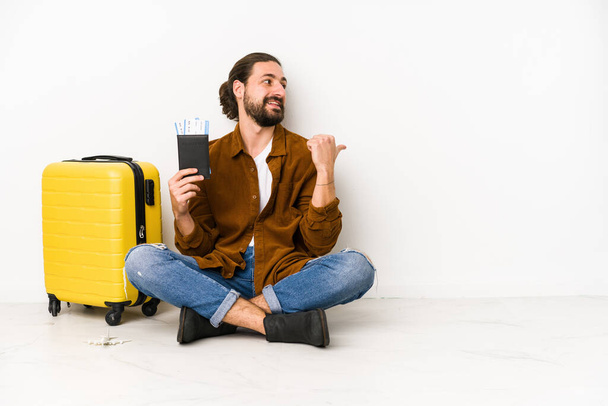 Young caucasian man sitting holding a passport and a suitcase isolated points with thumb finger away, laughing and carefree. - Foto, afbeelding