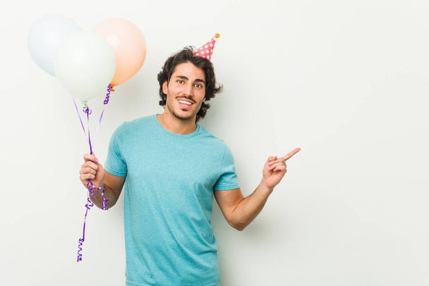 Partiyi kutlayan genç adam işaret parmağıyla mutlu bir şekilde gülümseyen balonlar tutuyor.. - Fotoğraf, Görsel