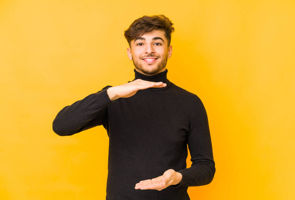 Jeune homme arabe isolé sur un fond jaune tenant quelque chose avec les deux mains, présentation du produit
. - Photo, image
