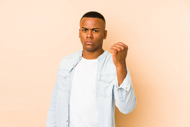 Young latin man isolated on beige background showing fist to camera, aggressive facial expression. - Photo, Image