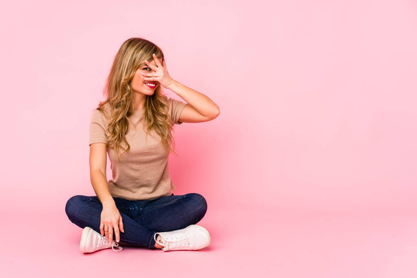 Νεαρή ξανθιά καυκάσια γυναίκα που κάθεται σε ένα ροζ στούντιο ανοιγοκλείνει τα μάτια της στην κάμερα με τα δάχτυλα, ντροπιασμένη να καλύπτει το πρόσωπο. - Φωτογραφία, εικόνα