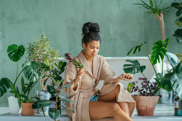 Pozitív kinézetű fekete hajú göndör afrikai többnemzetiségű nő ül előtt fürdőkád zöld fürdőszoba körül növények. Női higiénia, bolygómentés, környezetbarát, nulla hulladék, újrahasznosítási koncepció. - Fotó, kép