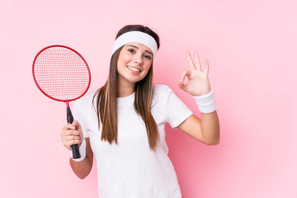 Junge kaukasische Frau spielt Badminton isoliert fröhlich und selbstbewusst zeigt ok Geste. - Foto, Bild