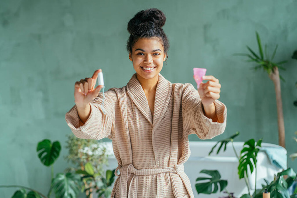 Çevre dostu Afro-Amerikalı kadın iki elinde tampon ve regl bardağı tutuyor, yan bakıyor, banyo içinde bej bornoz giyiyor, karşılaştırıyor. Regl, sıfır atık, yeniden kullanım kavramı. - Fotoğraf, Görsel