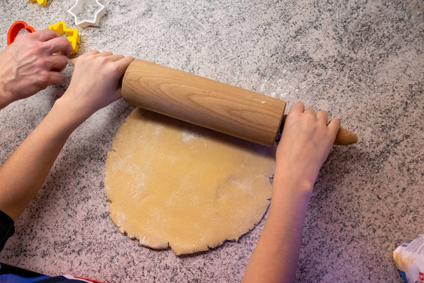Ребенок печет печенье с мамой или бабушкой, выкатывает тесто и использует формы, чтобы сделать вырезки печенья в рождественское время
 - Фото, изображение