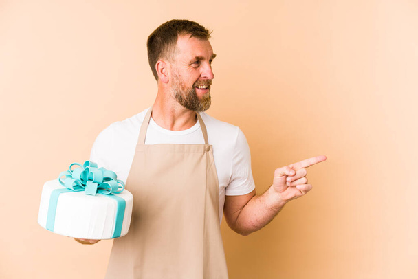 Senior holding a cake isolated on beige background smiling and pointing aside, showing something at blank space. - Foto, afbeelding