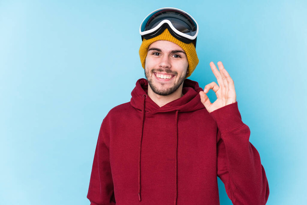 Jeune homme caucasien portant un vêtement de ski isolé gai et confiant montrant geste ok
. - Photo, image
