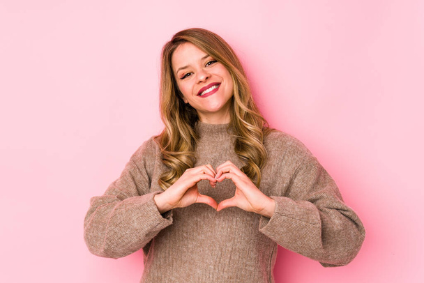 Young caucasian woman isolated on pink background smiling and showing a heart shape with hands. - Foto, Imagen