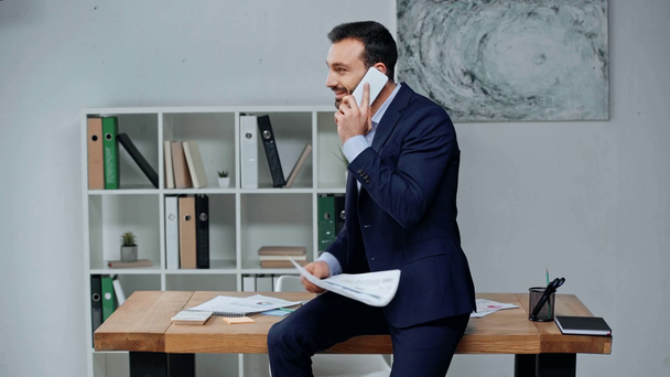 Lächelnder Geschäftsmann sitzt am Schreibtisch, hält Dokument in der Hand und spricht auf dem Smartphone - Filmmaterial, Video