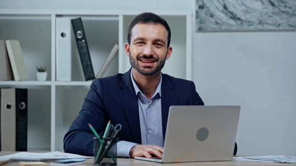 cheerful businessman typing on laptop and smiling at camera - Footage, Video