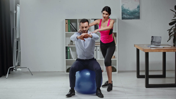 young trainer helping businessman with doing sit ups on fitness ball in office - Video, Çekim