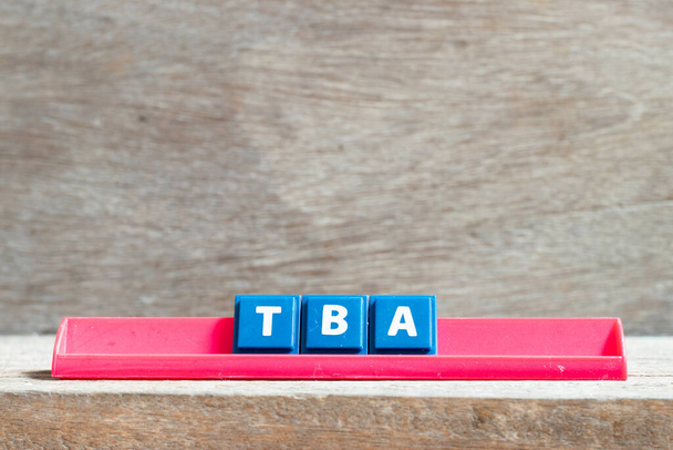 Επιστολή πλακιδίων σε κόκκινη σχάρα στη λέξη TBA (σύντμηση της οποίας πρέπει να ανακοινωθεί) σε φόντο ξύλου - Φωτογραφία, εικόνα