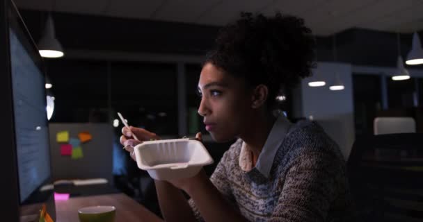 Vista laterale di una donna d'affari mista che lavora fino a tardi in un ufficio moderno, seduta alla scrivania a guardare lo schermo del computer, mangiare cibo da asporto
 - Filmati, video