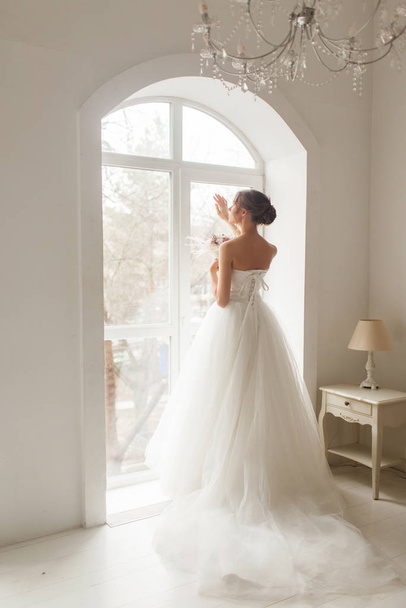 Joven novia en un hermoso vestido sosteniendo un ramo de flores posando cerca de la ventana en un estudio blanco brillante. Concepto de boda
. - Foto, Imagen