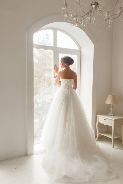 Young bride in a beautiful dress holding a bouquet of flowers posing near window in bright white studio. Wedding concept. - Foto, afbeelding
