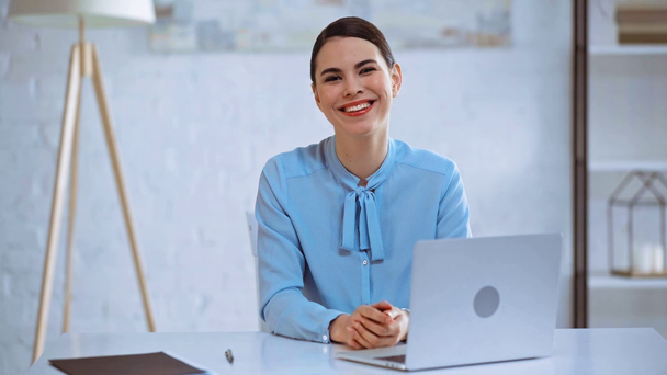 szczęśliwy businesswoman uśmiecha się w pobliżu laptopa w biurze  - Materiał filmowy, wideo