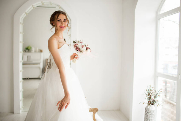 Young bride in a luxury dress holding a bouquet of flowers in bright white studio. Wedding fashion concept. - Foto, Imagem