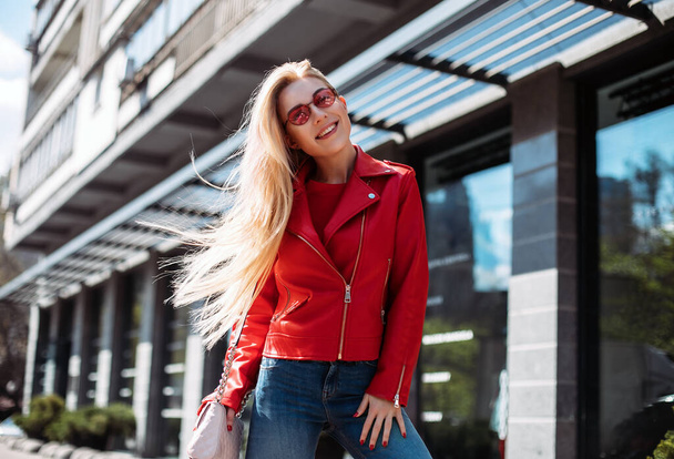 happy young stylish girl in leather jacket and jeans looking in camera walking around city street.beautiful blonde with long hair charmingly smiling. - Fotografie, Obrázek