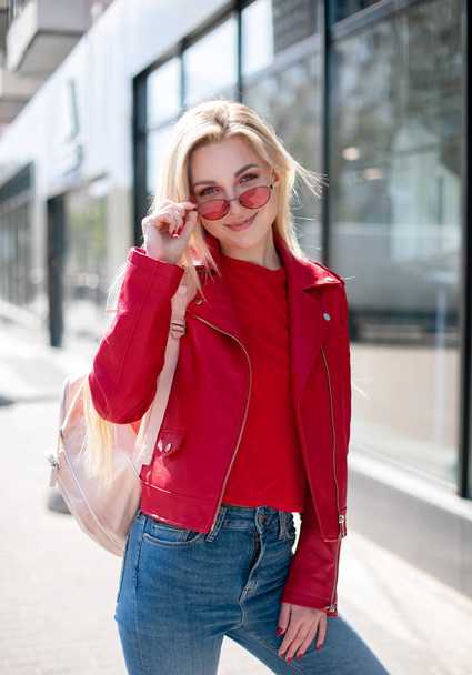 Fashion style portrait of young beautiful smiling young woman wearing a red leather jacket, posing at city street with magnificent architecture. Full body portrait. - Fotó, kép