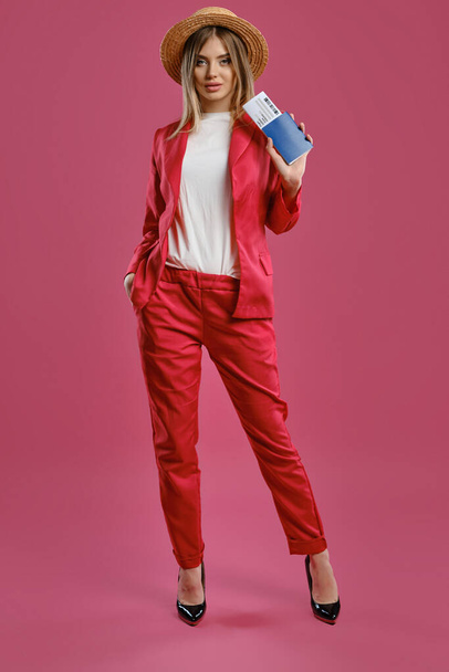 Blonde model in straw hat, white blouse, red pantsuit and high black heels. She holding passport and ticket, posing on pink background. Full length - Foto, imagen